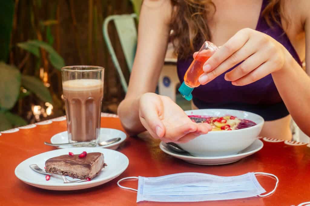 woman using antibacterial gel on hands before start to eating her healthy food in tropical asian cafe . smoothie bowl , raw cake and coffee on table with sanitizer on bottle