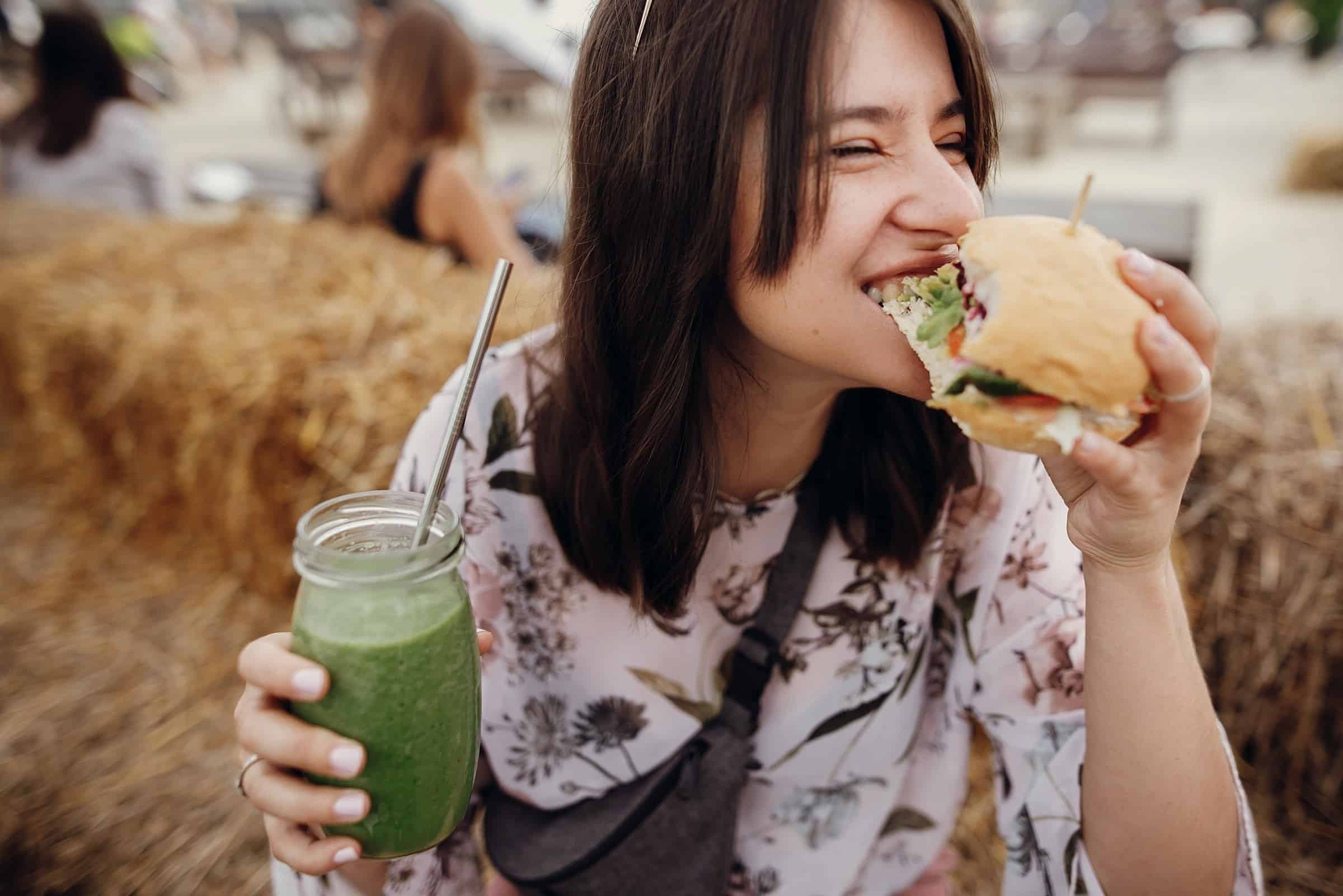 burger and holding smoothie in glass jar in hands at street food festival. Happy boho woman biting burger with drink in summer street